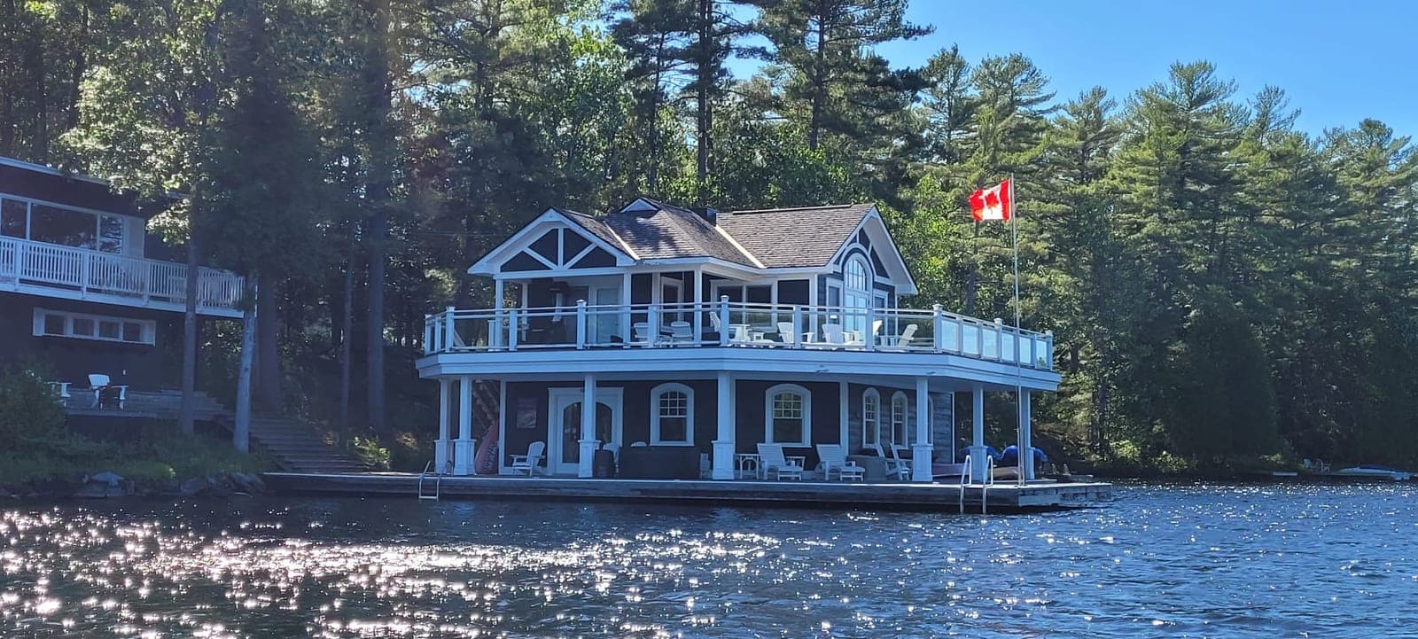 Two-storey boathouse with white columns, arched openings and a glass-railed upper deck, built out over open water below a cottage in the pines