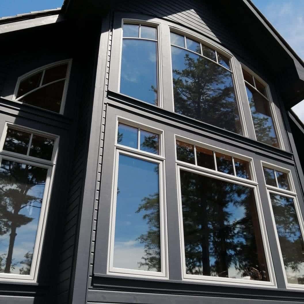 Close view of a gable window wall, arched multi-pane sashes with white trim against dark siding, pines reflected in the glass
