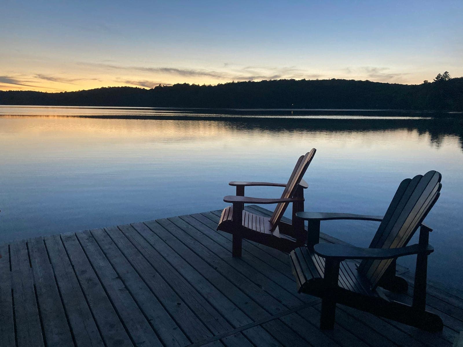 Two Adirondack chairs on a wooden dock at dusk with a calm lake and treed shoreline behind