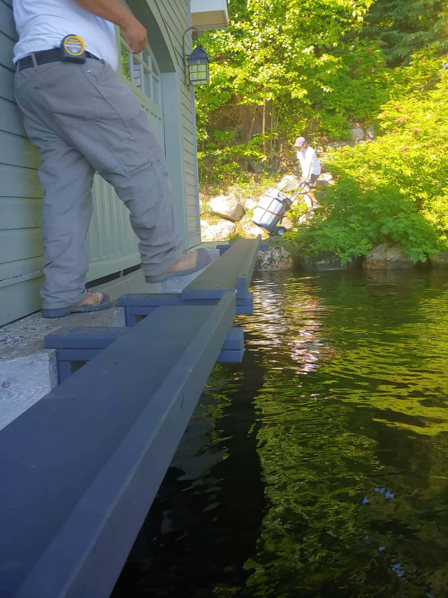 New dock framing set along the wall of a boathouse over clear water, one worker walking the fresh beams and another bringing gear down the shore