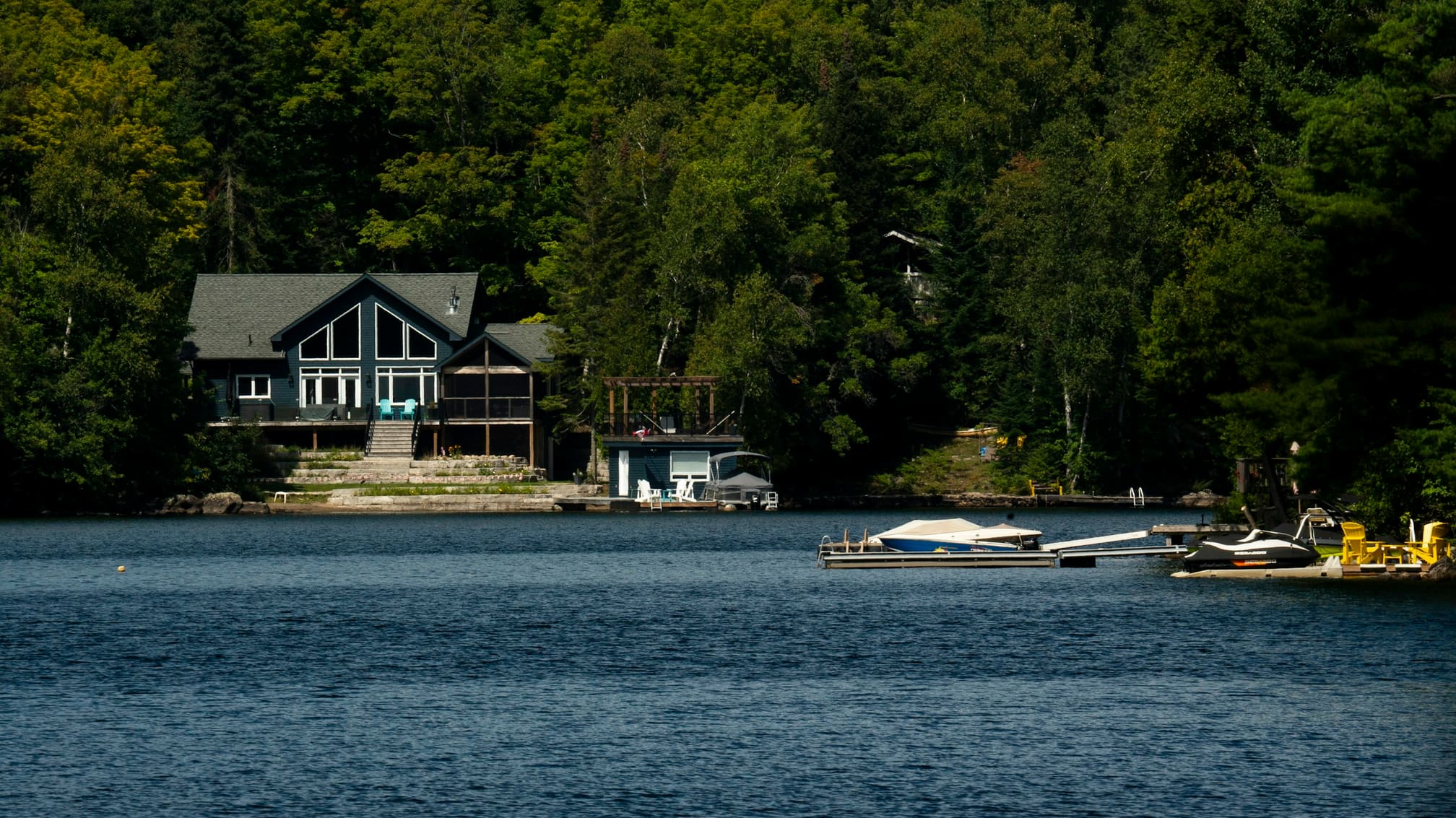 Dark-sided lakeside cottage with a boathouse and docks below on a treed Canadian lake shore