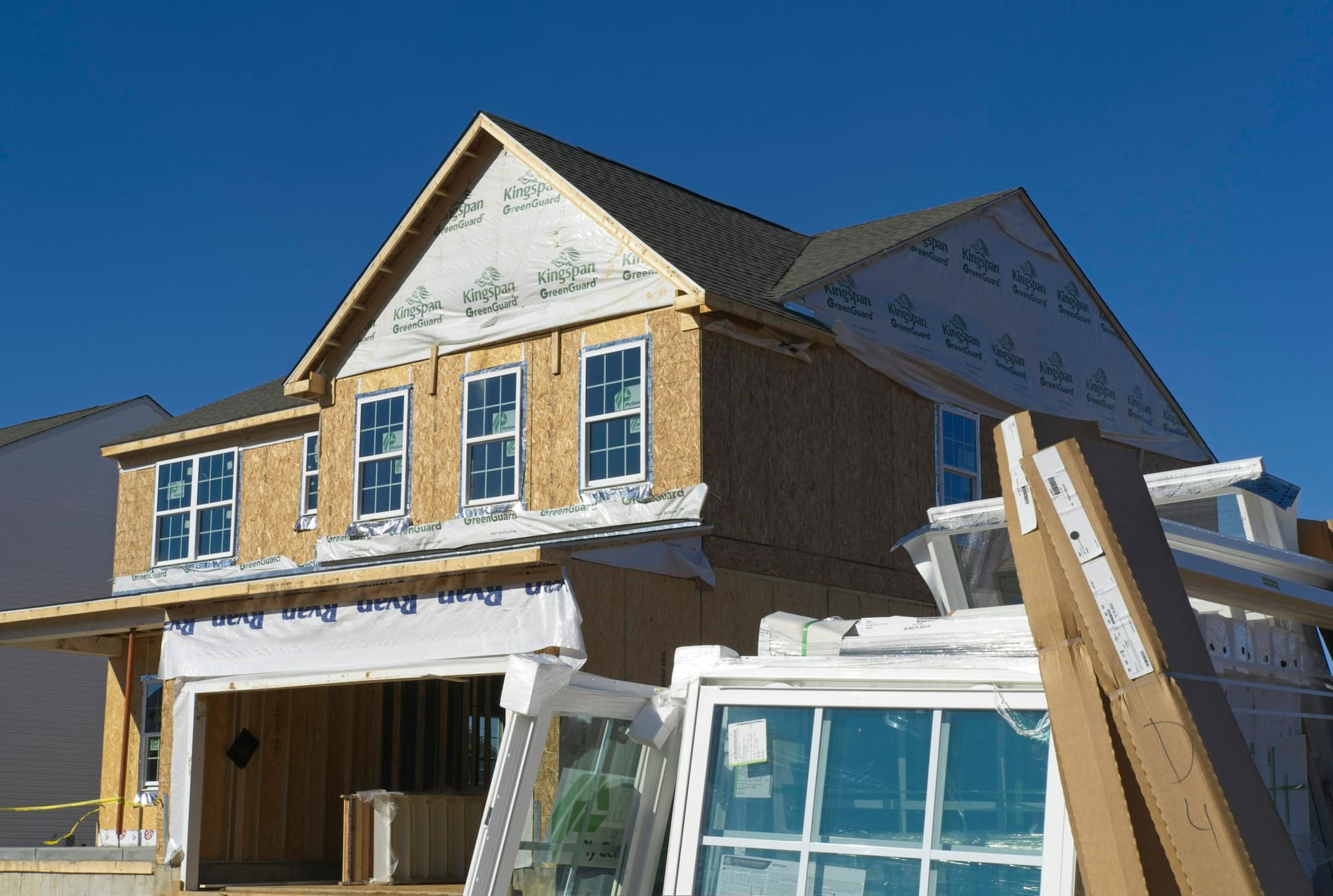 Two-storey house under construction with sheathing and house wrap on and windows waiting on site